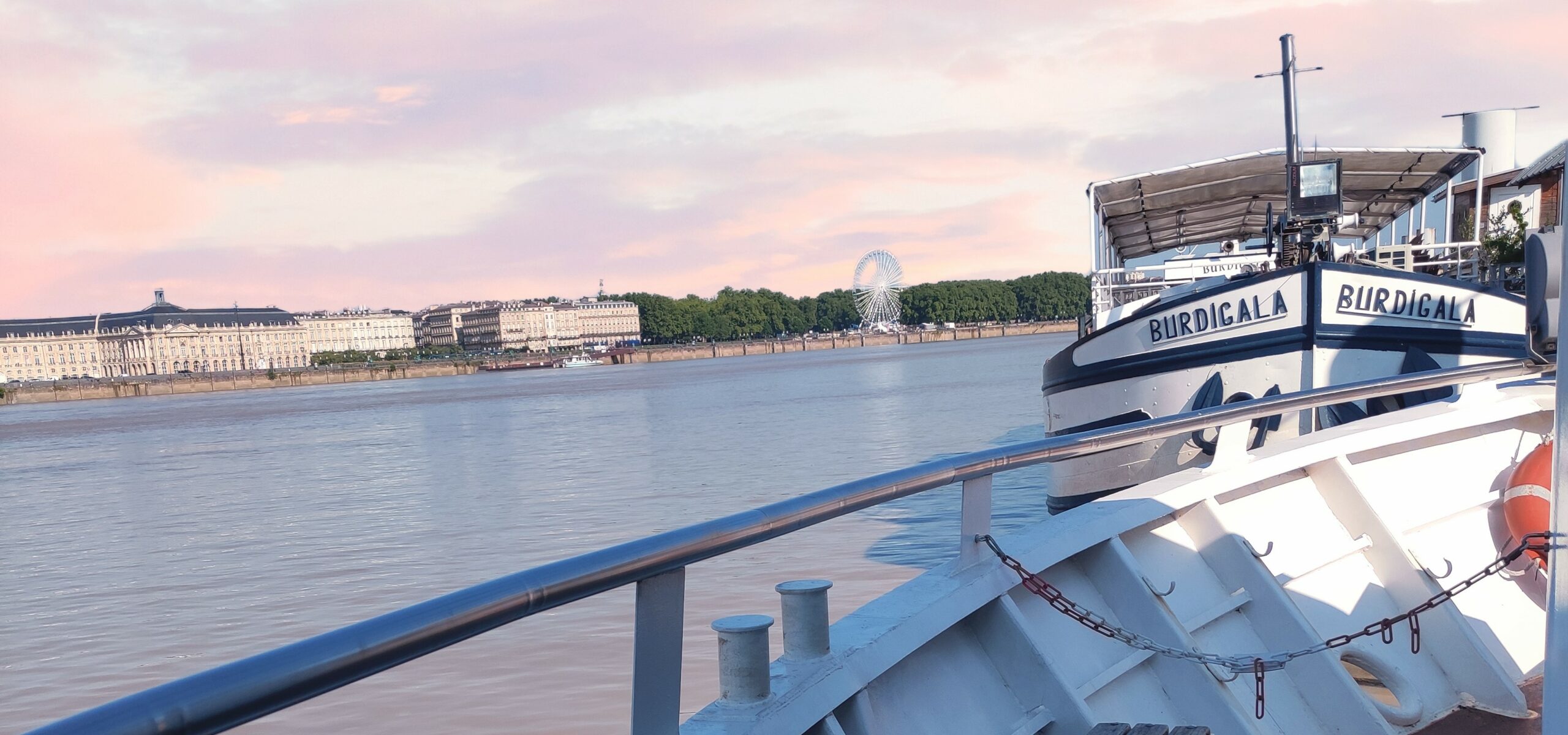 Photo prise depuis l'Aquitania amarré au ponton privé de Croisières Burdigala, face au Burdigala 1 au lever du soleil, avec en arrière-plan la Place des Quinconces, une grande roue et la Place de la Bourse.