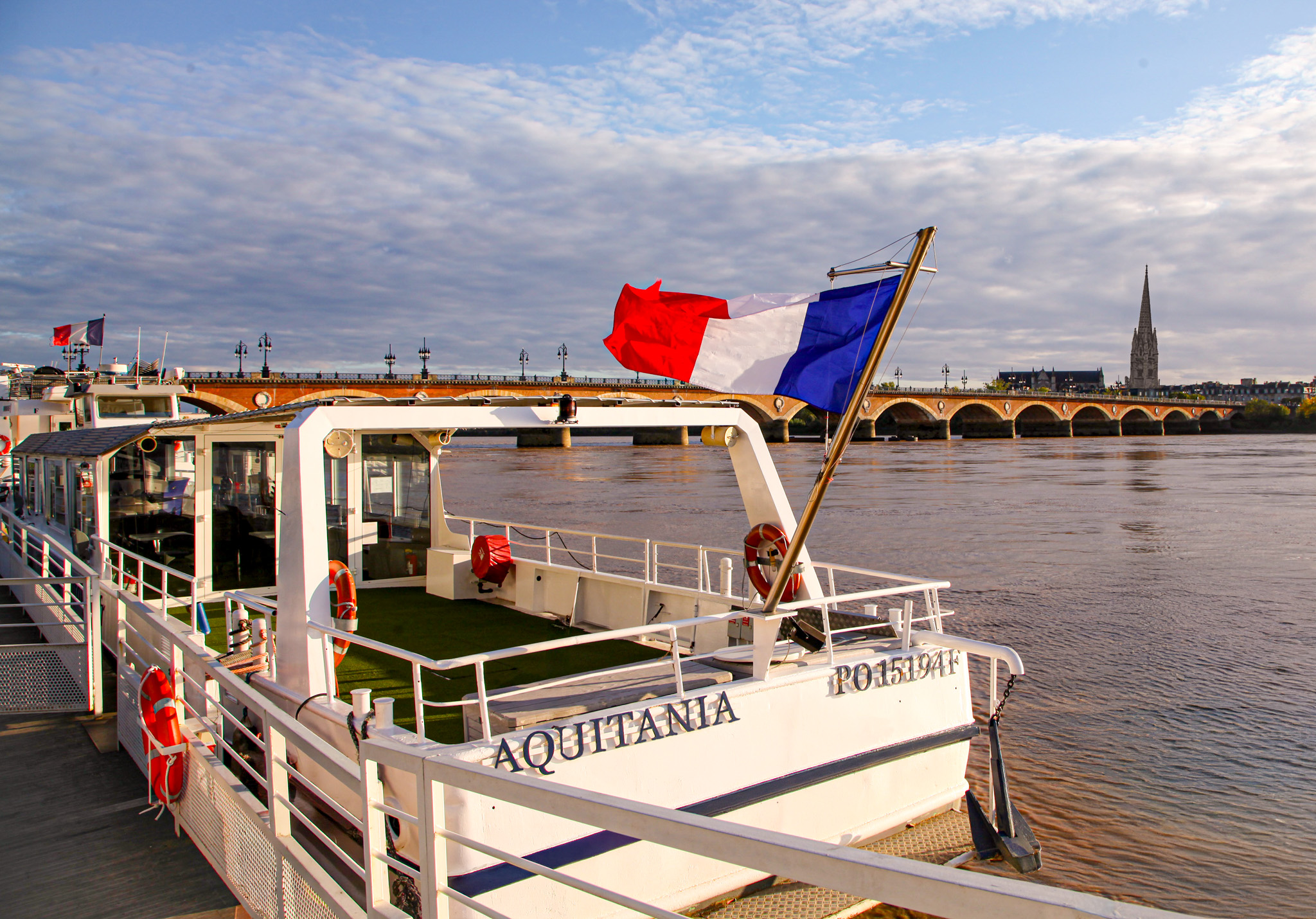 Vue depuis le ponton de Croisières Burdigala, mettant en avant l'Aquitania avec le drapeau français flottant au vent. En arrière-plan, le Pont de Pierre et la Basilique Saint-Michel de Bordeaux.