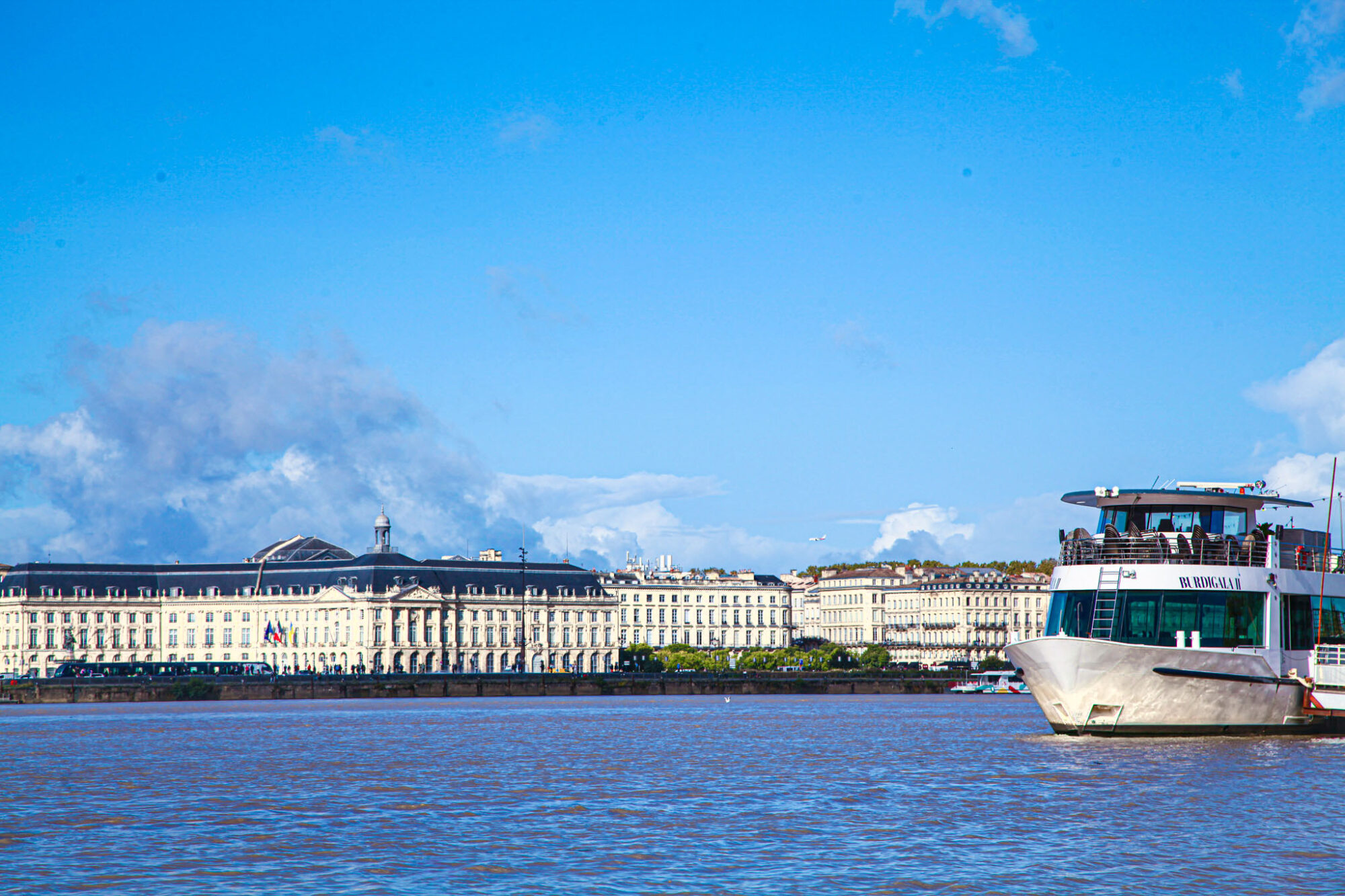 Le Burdigala 2 se dirige vers l'appareil photo, revenant d'une croisière sous un ciel bleu. En arrière-plan, la Place de la Bourse et le Miroir d'eau de Bordeaux.